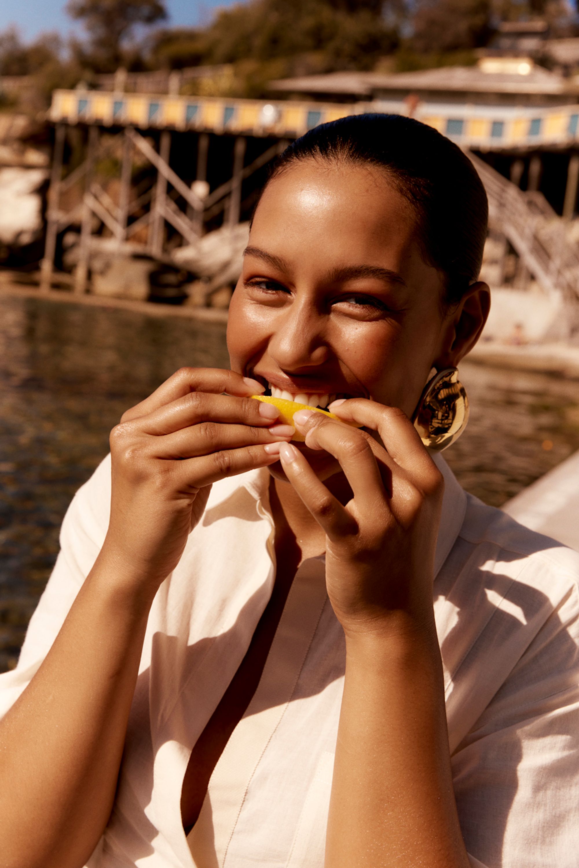 Portrait of a smiling woman eating a lemon slice outdoors near a wooden structure with a body of water and trees in the background.