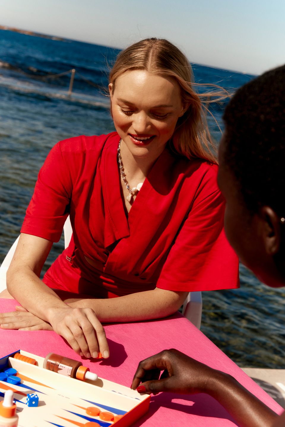 Two women play backgammon outdoors on a pink table near the ocean. One woman wears a red dress and a necklace.