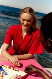 Woman in red top playing backgammon outdoors near the ocean for Mecca Summer Club 24 campaign