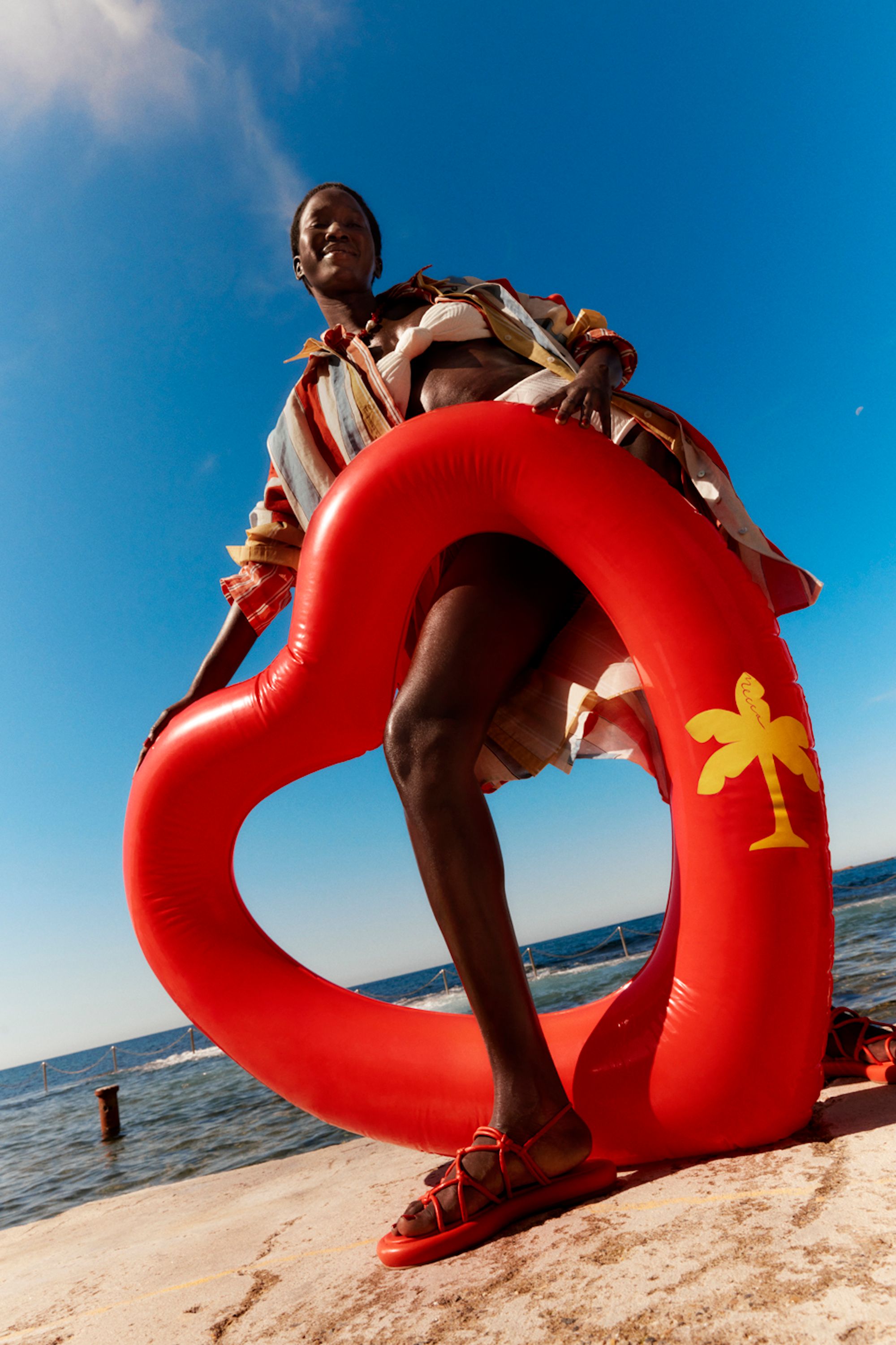 A young Black woman stands on the beach with a red heart-shaped flotation device under bright blue skies on a sunny day.