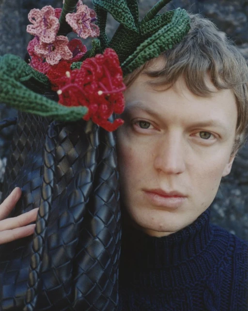 Headshot of a fair-skinned adult with a black woven purse and crocheted flowers against a weathered stone background. Fashion, beauty, and art.
