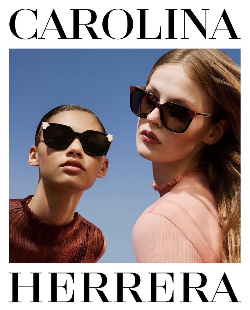 Two women model Carolina Herrera sunglasses against a blue sky. One wears a ribbed copper top and the other a sheer pink ruffled top.