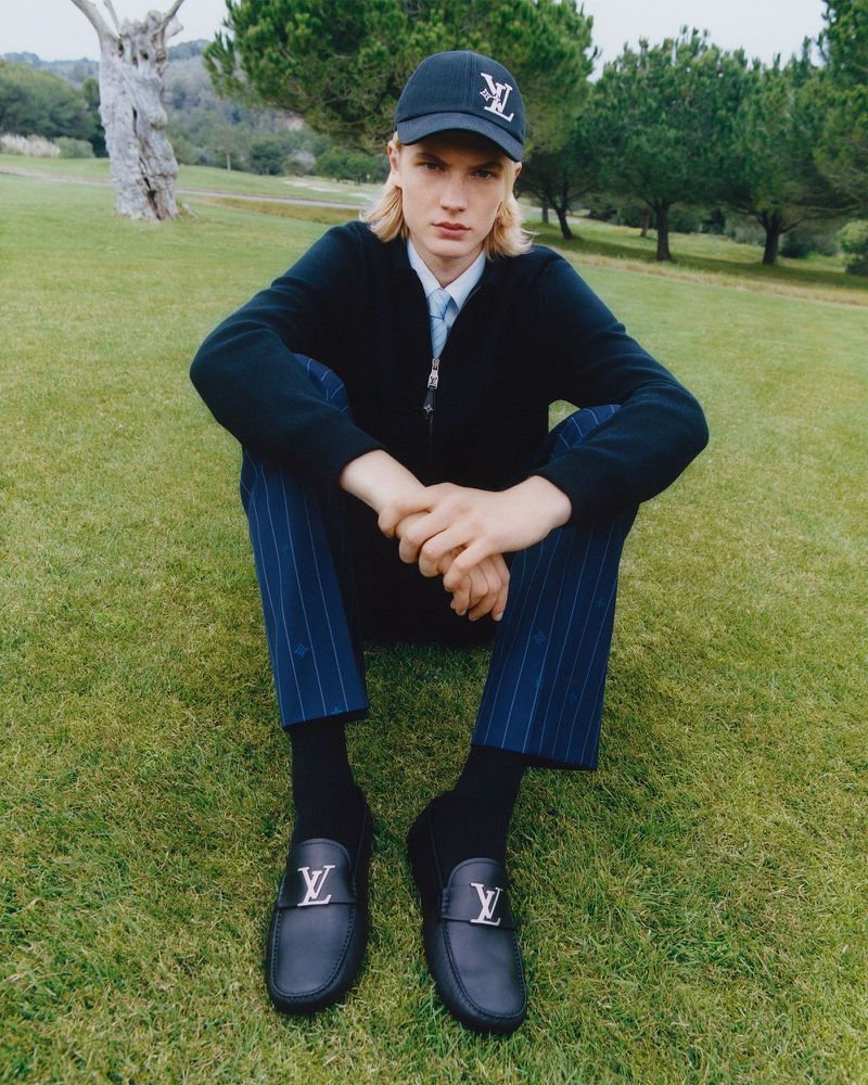 A stylish blond male model poses on a lush golf course wearing a navy Louis Vuitton cap, jacket, striped pants, socks, and loafers.
