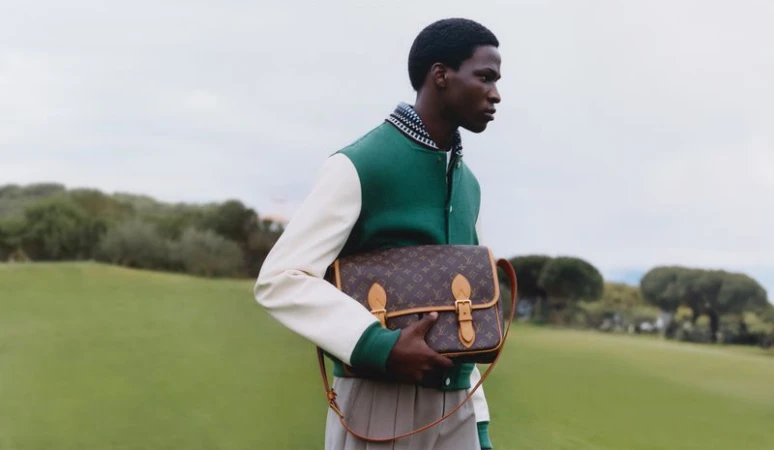 An adult male models a Louis Vuitton messenger bag in an outdoor field setting wearing a letterman jacket and khaki pants.