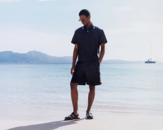 A dark-skinned male model stands on a beach in a printed button-down shirt, dark shorts, and sandals, with a sea and sky background.