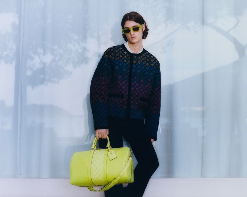 A person with sunglasses and a patterned sweater holding a lime duffel bag against a pale, textured wall.