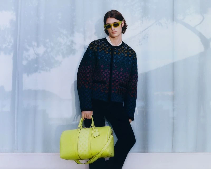 A person with sunglasses and a patterned sweater holding a lime duffel bag against a pale, textured wall.
