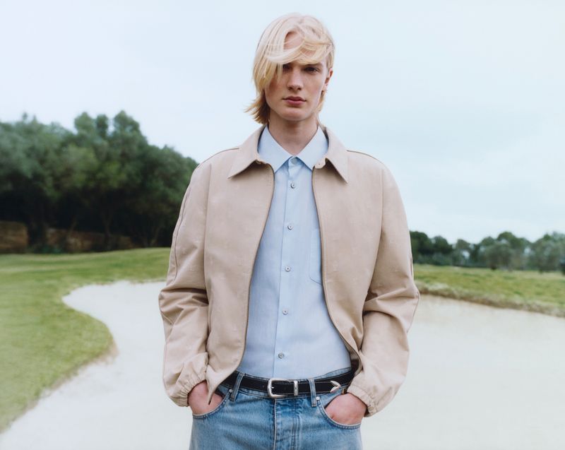 A young male model with blonde hair poses outdoors wearing a tan jacket, light blue shirt, blue jeans, and a black belt in a medium shot.