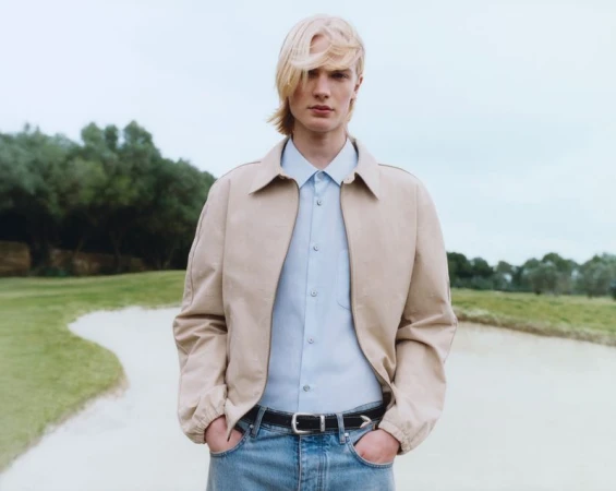 A young male model with blonde hair poses outdoors wearing a tan jacket, light blue shirt, blue jeans, and a black belt in a medium shot.