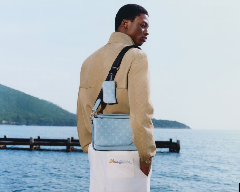 A tan-jacketed man with a blue Louis Vuitton bag stands outdoors with the ocean, a green mountain, and some pier posts in the background.
