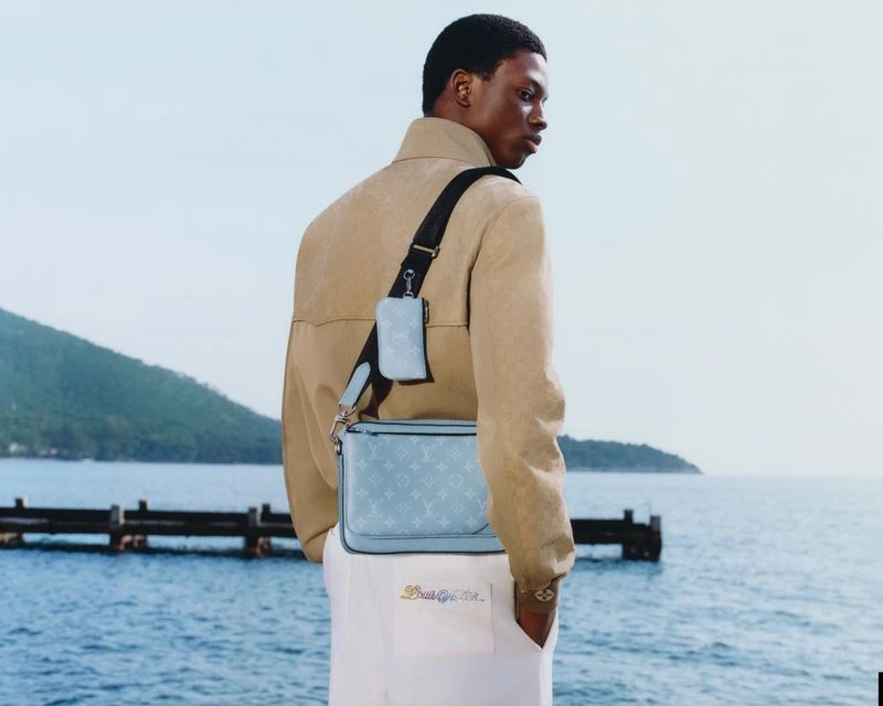 A tan-jacketed man with a blue Louis Vuitton bag stands outdoors with the ocean, a green mountain, and some pier posts in the background.