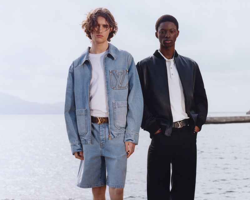 Two fashion models pose outdoors wearing denim and black leather jackets with LV logo belts, against a blurred sea and sky backdrop.