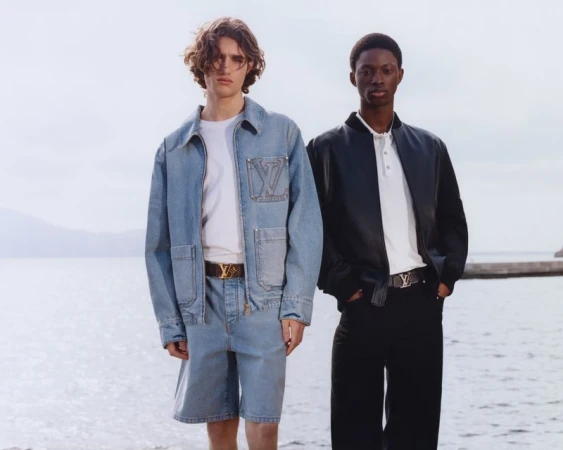 Two fashion models pose outdoors wearing denim and black leather jackets with LV logo belts, against a blurred sea and sky backdrop.