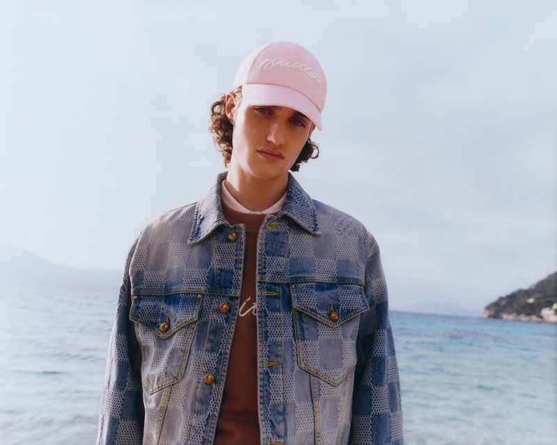 Young man wearing denim jacket and pink baseball cap poses against a coastal backdrop with blue ocean and pale sky on a sunny day.