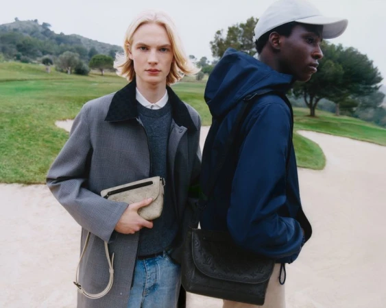 Two male models posing on a golf course wearing Louis Vuitton menswear, holding designer shoulder bags and jackets.