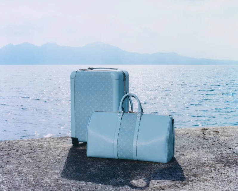 Light blue luggage set featuring a suitcase and duffel bag on a concrete surface with the sea and mountain range in the background.
