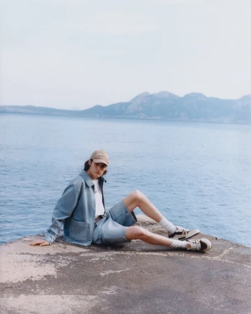 Man in denim jacket and shorts sits on concrete by the sea with mountains in the distance, wearing a baseball cap and sneakers.
