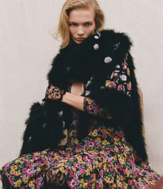 Blonde model in a floral dress and black faux fur stole, posing indoors against a neutral wall. Fashion photography, full-length.
