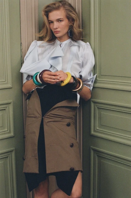 Blonde woman in a light blue blouse, black skirt, and tan trench coat worn as skirt, against an olive green door, accessorized with bracelets.