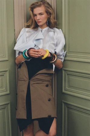 Blonde woman in a light blue blouse, black skirt, and tan trench coat worn as skirt, against an olive green door, accessorized with bracelets.