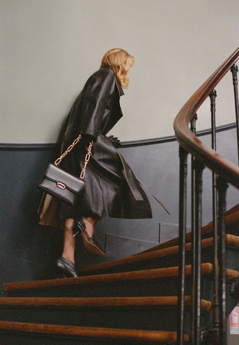Woman in a black leather trench coat and black heels climbs wooden stairs with a black purse and gold chain over her shoulder.