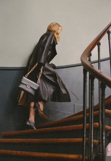 Woman in a black leather trench coat and black heels climbs wooden stairs with a black purse and gold chain over her shoulder.