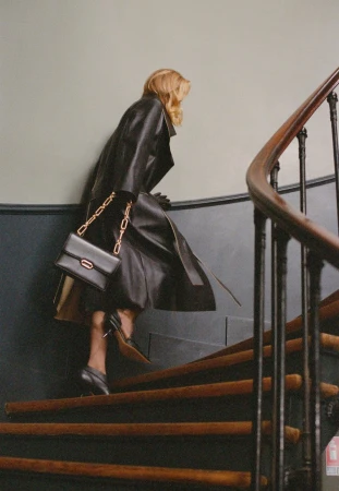 Woman in a black leather trench coat and black heels climbs wooden stairs with a black purse and gold chain over her shoulder.