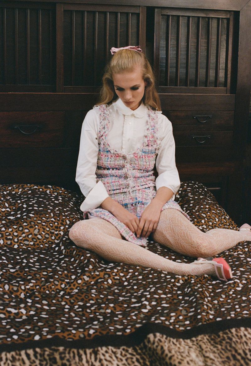A young blonde woman in a sleeveless dress sits cross-legged on a bed with an animal print blanket in an indoor setting with wooden furniture.