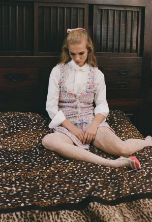 A young blonde woman in a sleeveless dress sits cross-legged on a bed with an animal print blanket in an indoor setting with wooden furniture.