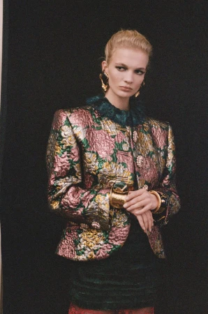 Model wearing a patterned metallic floral jacquard jacket and statement gold earrings against a dark background.