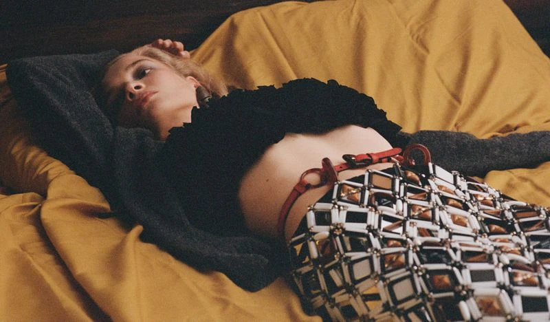 A young adult woman lies on a bed with a yellow blanket wearing a black top, gray cardigan and an elaborate metallic skirt.