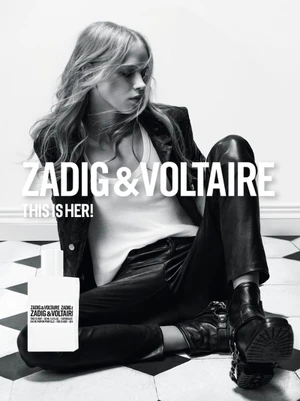 Model seated on a black and white tiled floor wearing a leather jacket and pants with a perfume bottle in the foreground.