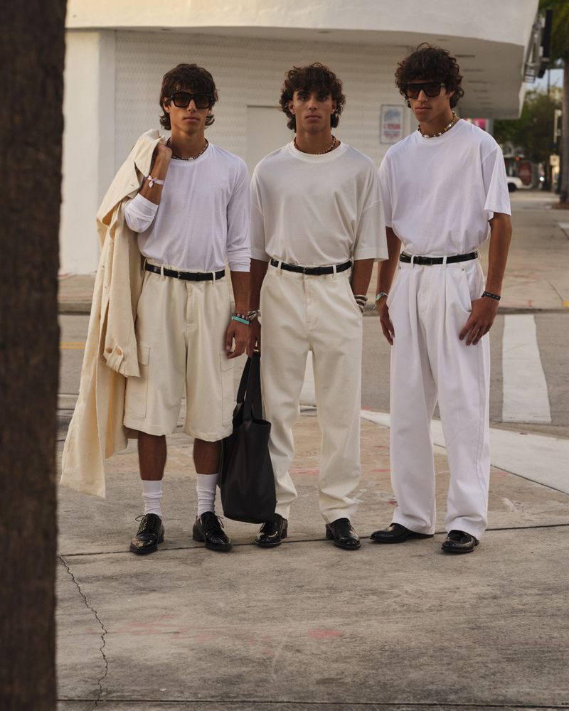 Three men pose on a crosswalk in matching outfits, wearing sunglasses, white shirts, and pants, with one holding a tan coat.