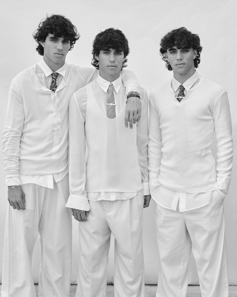 Three young adult males stand in a neutral environment, all wearing white shirts and trousers, with two wearing ties in a black and white portrait.