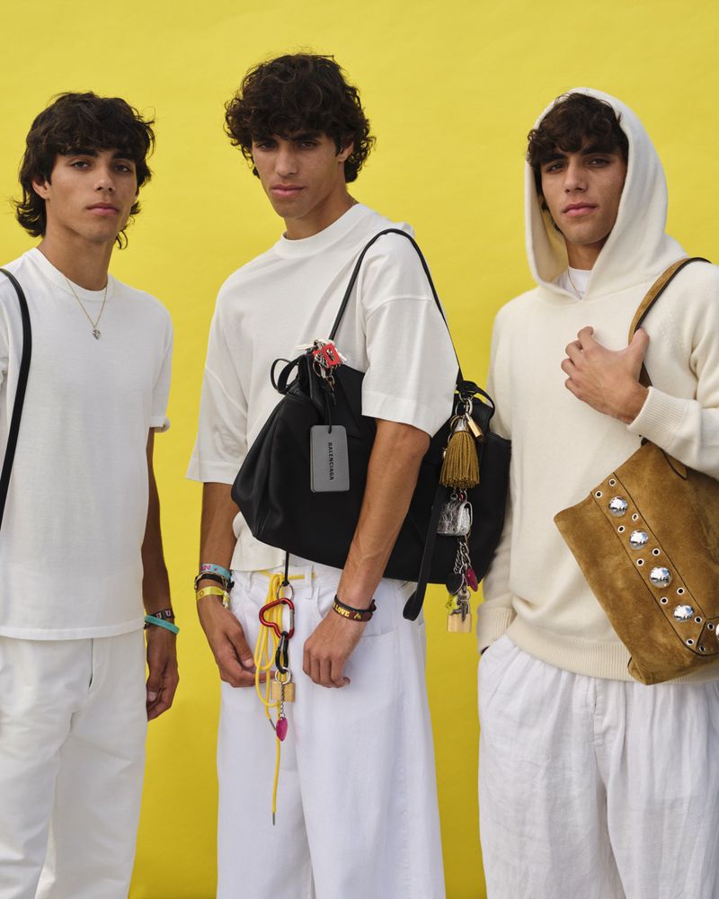 Three young adult men pose in a medium shot against a bright yellow background. They are wearing casual clothes and carrying shoulder bags.