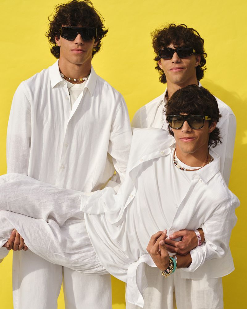 Three young men pose against a yellow background, one being held in arms. They wear white shirts, pants, and sunglasses, completing the fashion shot.