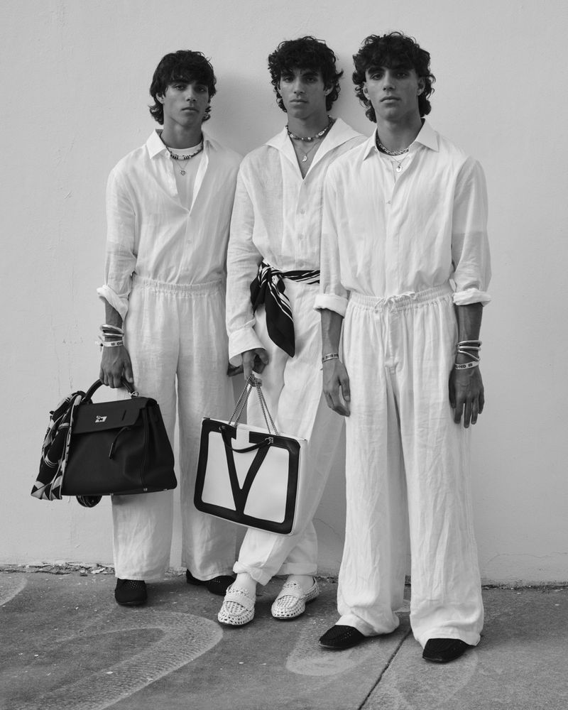 Three male fashion models pose in matching white linen outfits with handbags against a neutral wall in a black and white photograph.