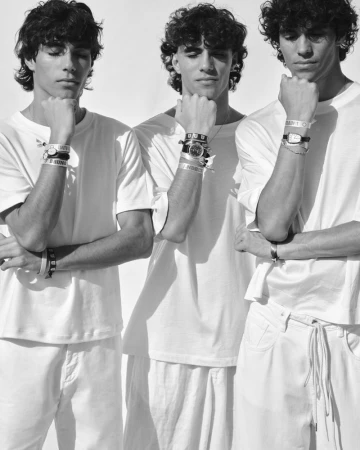 Three young adult males pose indoors in white T-shirts and pants, displaying bracelets on their wrists in a black and white fashion editorial.