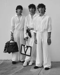 Three young men in white linen shirts and trousers holding designer handbags against a plain light wall in a monochrome portrait.