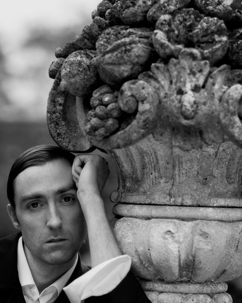 Monochrome portrait of a man in a suit leaning against an ornate stone vase with a floral arrangement, captured in soft, diffused light.