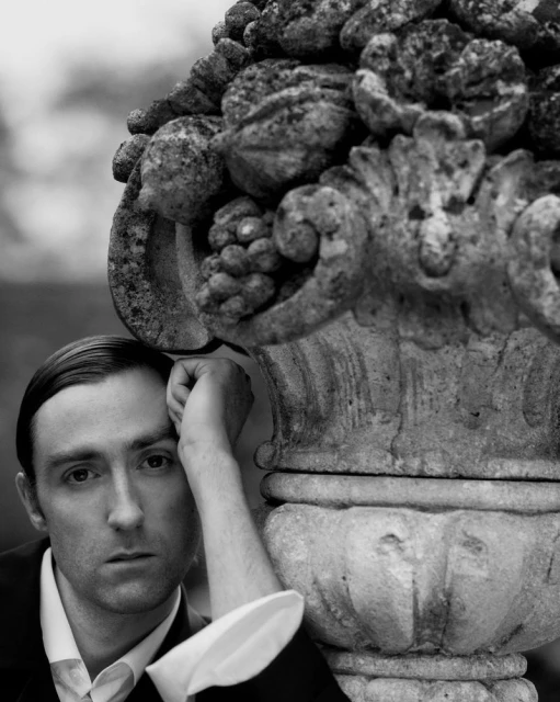 Monochrome portrait of a man in a suit leaning against an ornate stone vase with a floral arrangement, captured in soft, diffused light.