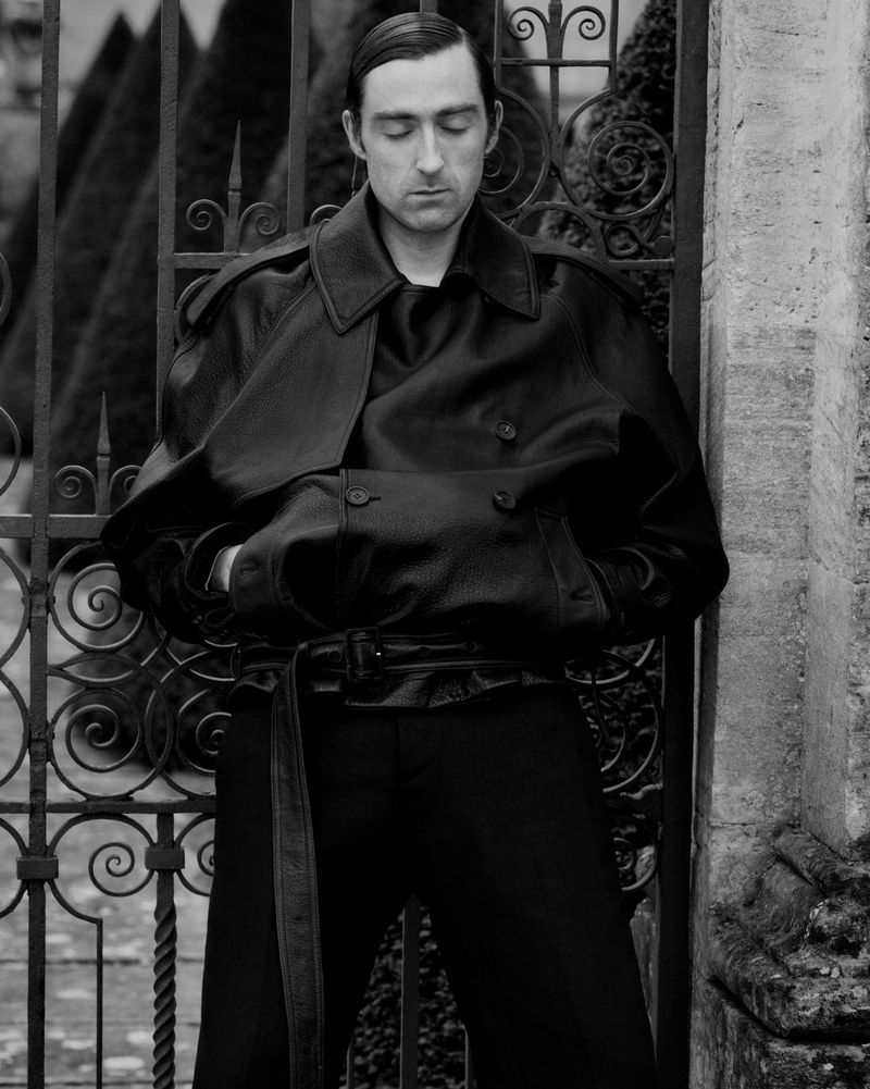Man in a black leather trench coat with a belt stands outdoors against an ornate metal gate and stone wall, posing for a fashion shot.