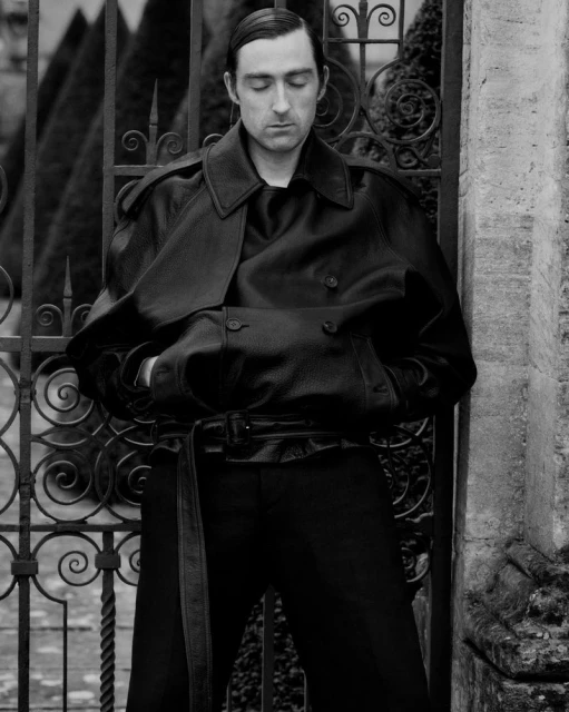 Man in a black leather trench coat with a belt stands outdoors against an ornate metal gate and stone wall, posing for a fashion shot.