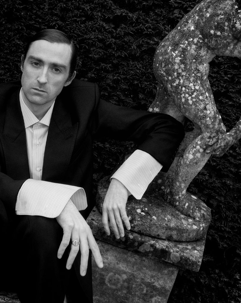 Man in dark suit with wide striped cuffs leaning against a weathered stone garden statue in black and white