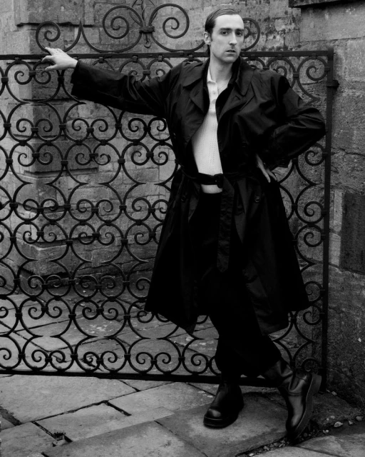 Monochrome full shot of a male model in a black trench coat posing against an ornate gate outdoors on a paved surface.