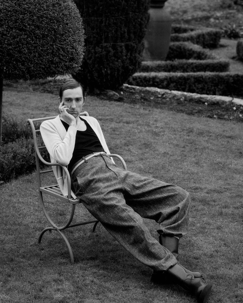 Black and white portrait of a seated adult male in outdoor garden, styled with black tank top, oversized patterned pants, boots and cardigan.
