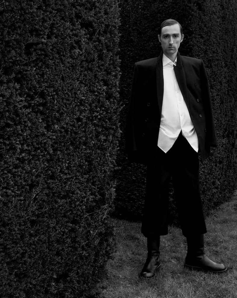 An adult male in a black suit and unbuttoned white shirt stands outdoors next to a tall hedge in a black-and-white fashion shot.