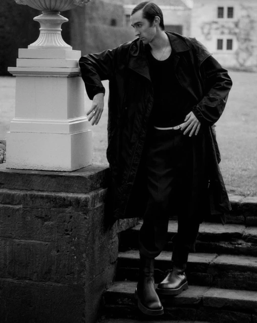 Monochrome full shot of an adult man in a trench coat standing on stone steps in an outdoor environment with grass and a building.