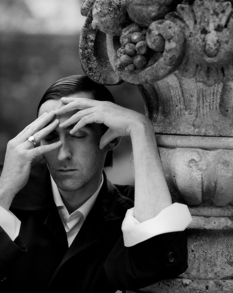 Monochrome portrait of a contemplative adult male in dark coat with eyes closed, hands on face, standing beside a weathered stone sculpture.