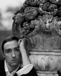 Black and white portrait of a man resting his head against a weathered stone urn outdoor.